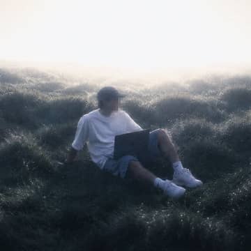 A figure with a laptop sitting in misty grass, face pixelated